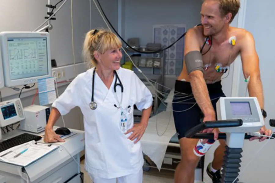 Bildet viser Turid Appeland som står ved siden av en pasient som sitter på en ergometersykkel