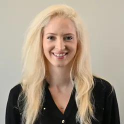 Elisabeth Askimdal Hersvik, iført en svart skjorte med små dekorative detaljer, står foran en ensfarget, lys bakgrunn. Hun har langt, lyst hår.