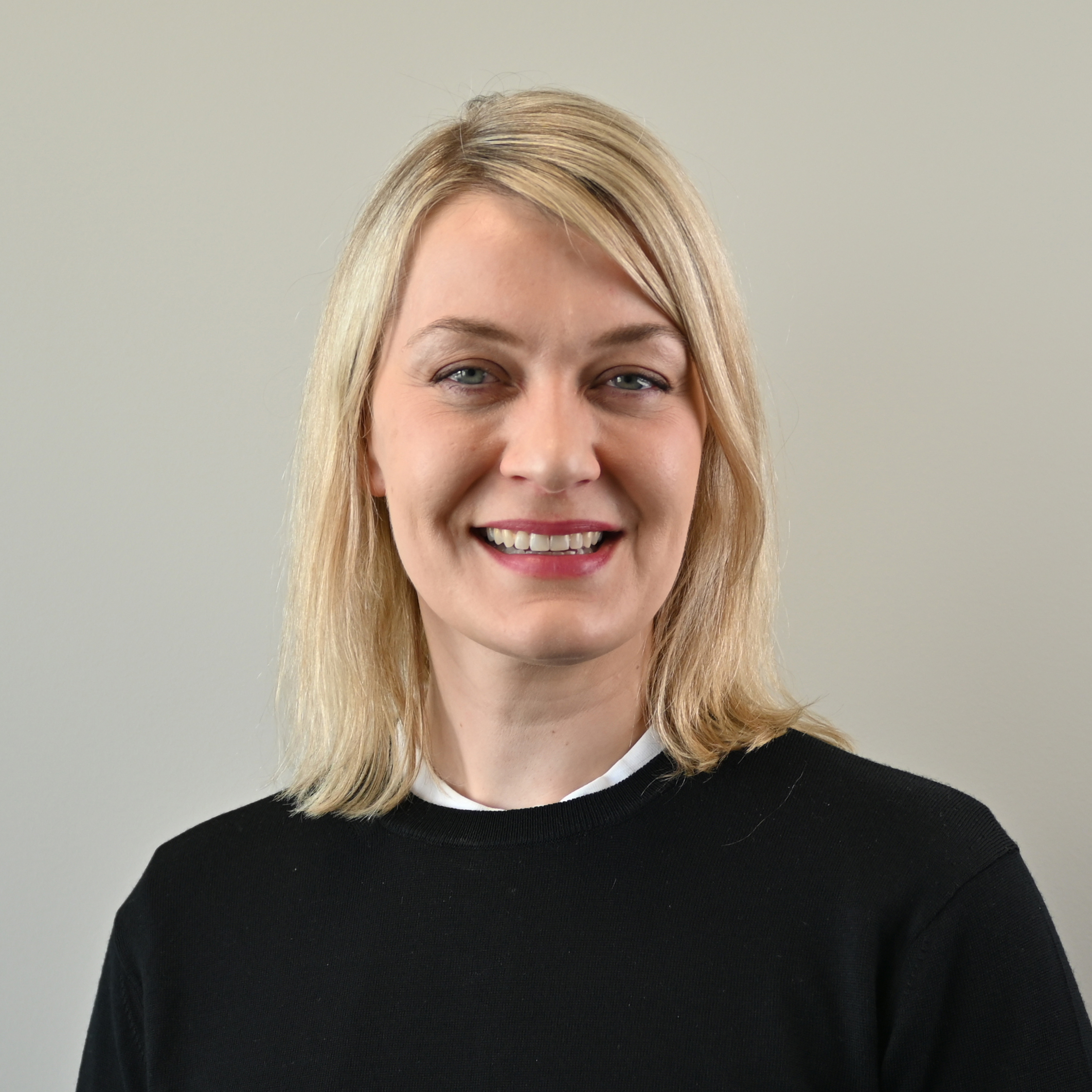 Hanna Høyem Aronsen, iført en svart genser over en hvit skjorte, står foran en ensfarget, lys bakgrunn. Hun har skulderlangt, lyst hår.