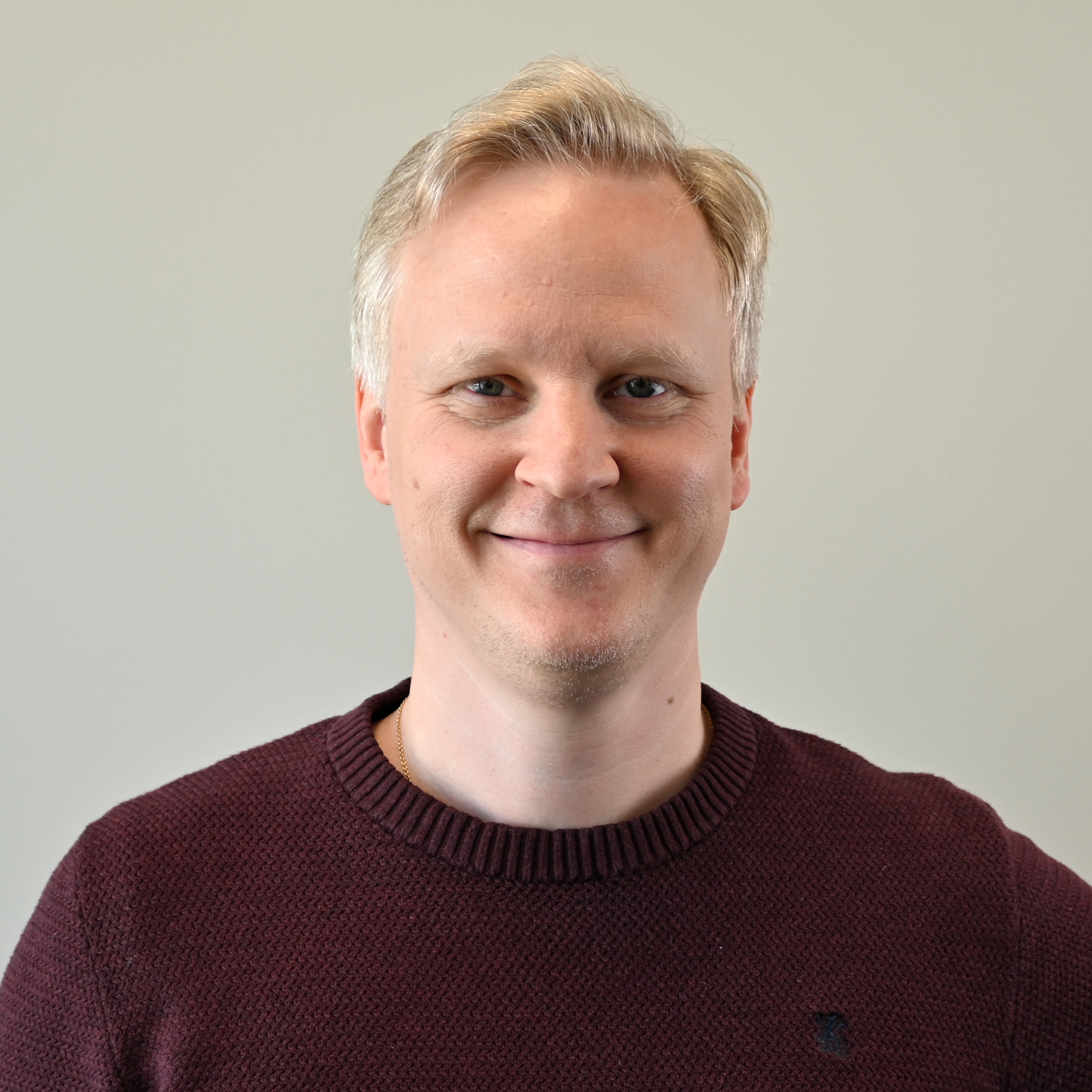 Jonas Vardal, iført en mørk burgunderrød strikkegenser, står foran en ensfarget, lys bakgrunn. Han har kort, lyst hår