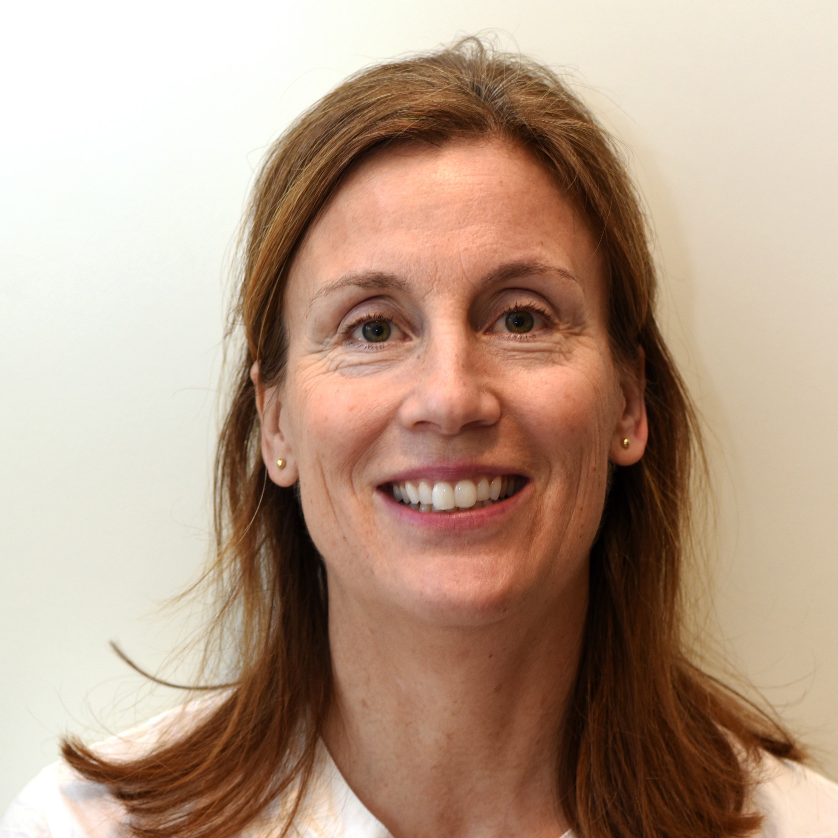 Gudrun Breistøl, med skulderlangt, brunt hår, iført en hvit topp og små øredobber, står foran en ensfarget, lys bakgrunn.