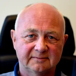 Rune Kløvtveit, med kort, grått hår, iført en blå poloskjorte, sitter foran en mørk stol med lys bakgrunn.